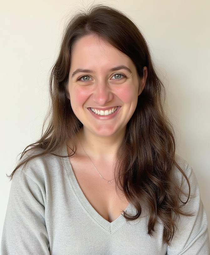 A professional portrait of Katie wearing a soft gray V-neck sweater and wavy brown hair loose over her shoulder, smiling directly at the camera.
